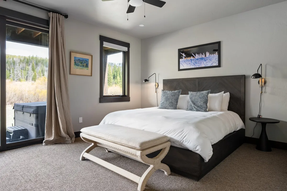 A serene bedroom with a comfortable bed adorned with decorative pillows, a large window providing outdoor views, artwork on the wall, and a stylish nightstand with a lamp. The space looks bright, tranquil, and designed for restful retreats.