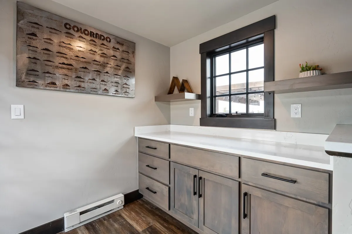 A cozy, modern kitchen interior with wooden cabinetry, a window framing picturesque mountain views, and Colorado mountain-themed art. The space combines functionality for cooking and working with an inviting ambiance and a seamless visual connection to the outdoors.