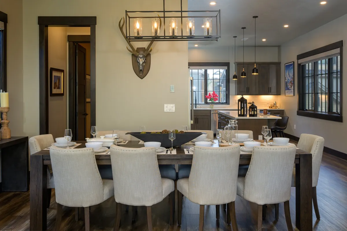 A spacious dining area with a large wood table, elegant chandelier, antler decor, and a view into the adjoining kitchen area. The room is set for family dinners or celebratory feasts, radiating warmth and sophistication.