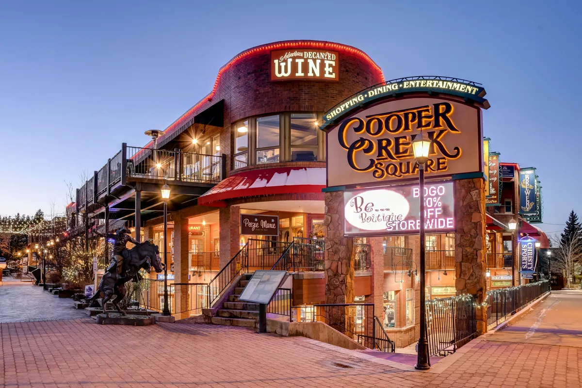 An exterior image showcasing the vibrant Cooper Creek Square, complete with a prominent sign, a wine shop above, an outdoor dining area, and a horse sculpture. The scene buzzes with activity, hinting at shopping, dining, and entertainment opportunities.