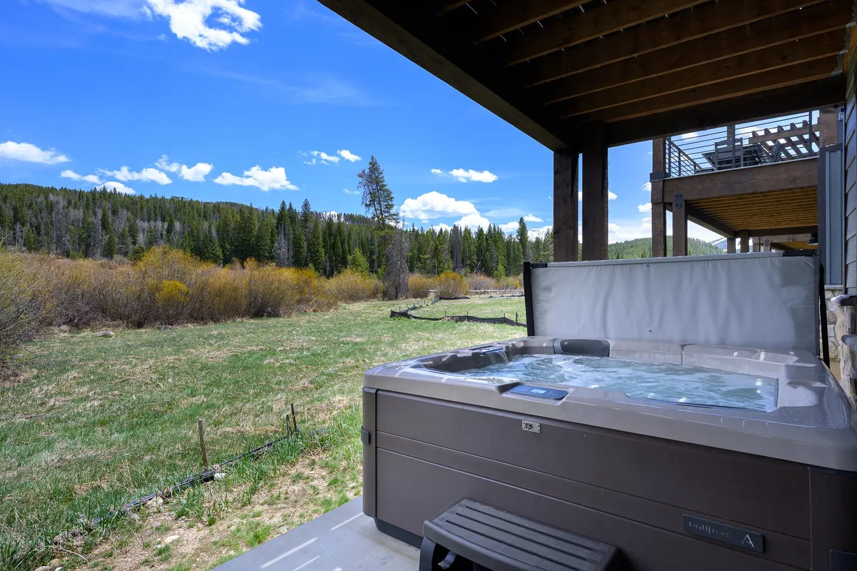 A luxurious outdoor hot tub sits on a private deck, overlooking a stunning natural landscape with mountains in the distance. The scene is tranquil, with cozy seating nearby and open sky above, suggesting relaxation while immersed in nature.
