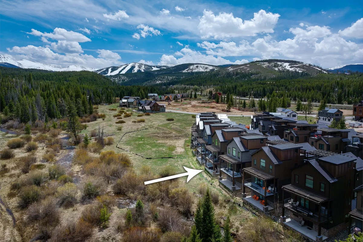 A panoramic view of a majestic mountain range in the background, framed by a row of houses and an open green space in the foreground. The setting evokes outdoor adventure and a tranquil atmosphere ideal for midday hikes or leisurely exploration.
