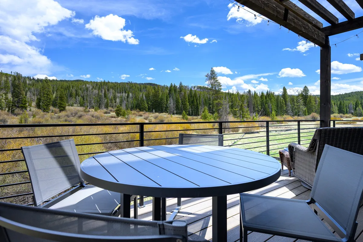 An inviting outdoor dining area under a wooden pergola, featuring a spacious table set for meals. The setting opens up to a lush surrounding landscape, ideal for al fresco dining, relaxing, and enjoying the tranquility of nature.
