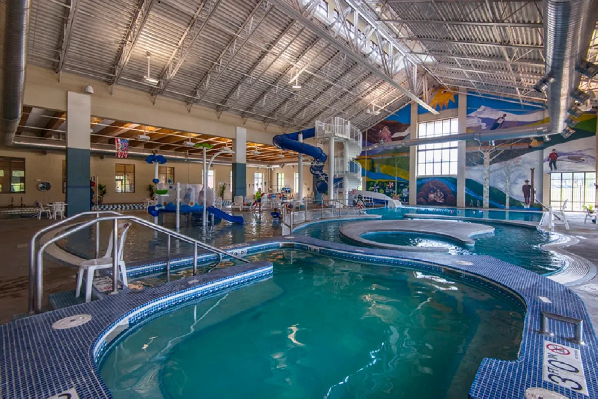 A vibrant, playful view of an indoor rec center pool and water slide with children playing, decorative murals, and an inviting atmosphere for family fun.